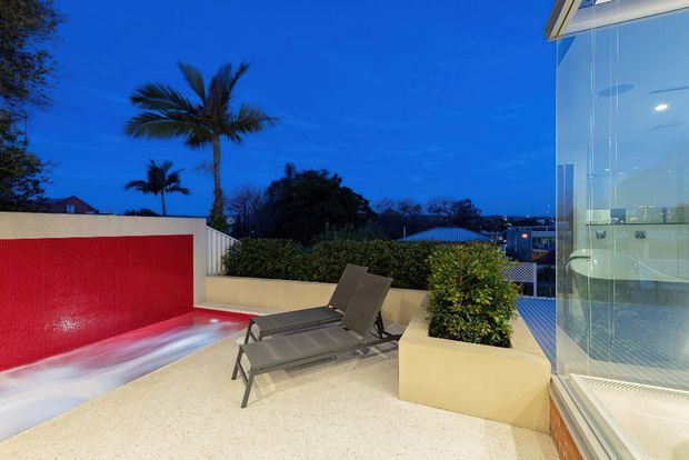 An evening exterior terrace beside a glazed modern building, featuring two reclining lounge chairs, pale paved flooring, raised cream-coloured planters with shrubs, and a vivid red tiled or rendered feature wall with integrated linear lighting. Tall palm trees and neighbouring rooftops are visible beyond the terrace, while the glass facade reveals a brightly lit interior bathroom or living space.