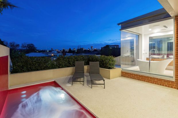 Twilight view of a contemporary rooftop terrace with a small illuminated pool or spa in the foreground, two lounge chairs, raised planter beds, and a glass-enclosed bright interior beyond. The terrace overlooks an urban neighbourhood, with clean geometric lines, pale paving, white walls, and a brick-clad base.