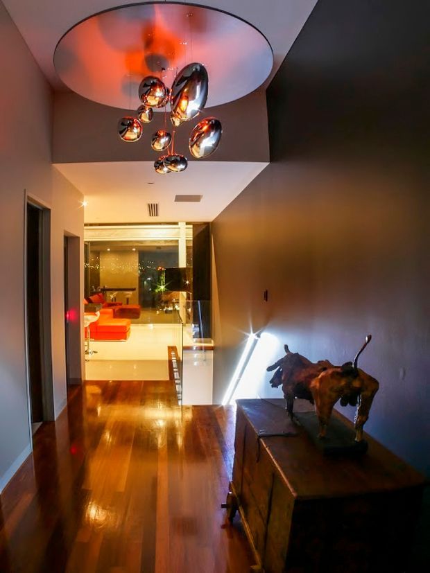 A dramatic contemporary residential hallway viewed toward a brighter living area. The corridor has glossy dark timber flooring, dark painted walls, a white ceiling with a large circular recessed feature, and a sculptural cluster of reflective pendant lights. A dark timber console with a decorative sculpture occupies the right foreground, while a vivid orange seating area is visible beyond a glass opening.