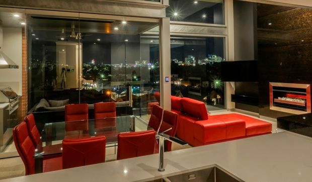 A sophisticated open-plan living and dining area at night, with floor-to-ceiling glazing overlooking city lights. The room features vivid red leather seating and dining chairs, a glass dining table, a large wall-mounted television, and a built-in fireplace set into a dark textured feature wall. A pale stone kitchen island or counter occupies the foreground, with recessed ceiling lighting and reflective surfaces creating a polished, contemporary atmosphere.