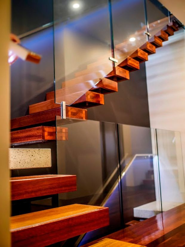 Close-up architectural view of a modern floating staircase with richly stained timber treads cantilevered from a dark wall. Clear glass balustrade panels are held by metal fittings, creating a dramatic open stairwell with reflective surfaces and integrated lighting. A small light-coloured stone or concrete accent block is visible within the stair structure.