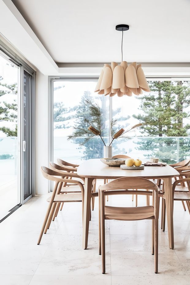 Bright dining area with a light timber dining table and matching curved chairs, positioned beside expansive floor-to-ceiling glazing. A sculptural natural-toned pendant hangs above the table, while white finishes and pale stone-look flooring create an airy, refined interior with views of trees and water beyond the glass.