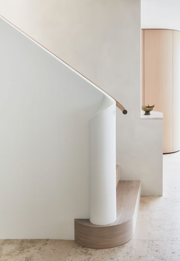 Minimalist interior view of a sculptural curved staircase with a smooth white cylindrical newel or structural element, pale timber handrail, and integrated pale timber steps. The space uses soft off-white plastered walls, light timber detailing, and a pale stone or terrazzo-like floor. A curved timber screen and small recessed display shelf are visible beyond the stair.
