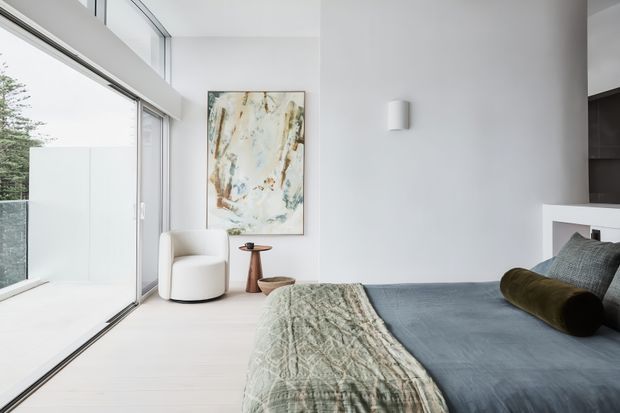 Bright contemporary bedroom with a low platform bed, blue-grey bedding, textured throw, and bolster cushions. The room features expansive floor-to-ceiling glazing, white walls and pale flooring, a small timber side table, abstract artwork, and a minimalist wall light. A dark built-in joinery element is partially visible behind the bed.