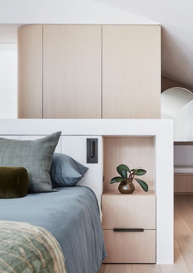 A softly lit contemporary bedroom corner featuring integrated pale timber cabinetry, a built-in bedside niche with a potted plant, and a bed dressed in muted blue-grey bedding with layered cushions. White architectural framing, subtle vertical timber grain, black hardware, and a curved mirror visible in the background create a calm, refined built-in joinery composition.