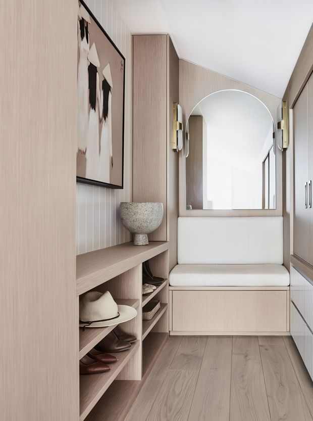 A refined built-in entryway or hallway with pale timber-look wall panelling, integrated open shelving, cabinetry, and a cushioned bench seat. An arched mirror is framed by wall sconces, while a neutral abstract artwork and carefully arranged accessories add understated decoration. The space has a narrow, compact footprint, light timber flooring, and a soft neutral palette.