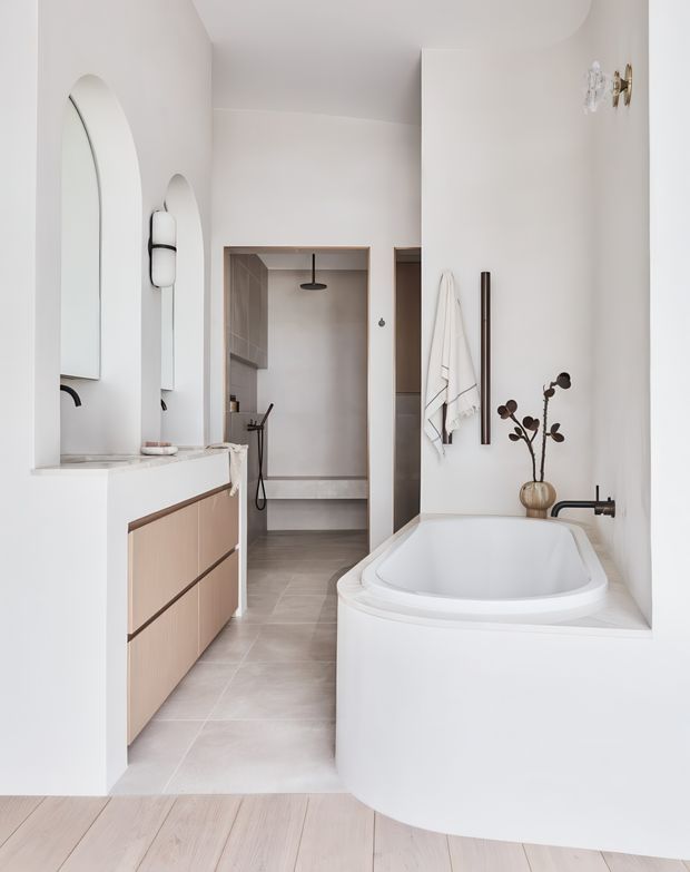 Bright, refined bathroom with a freestanding inset-style bathtub, walk-in shower, pale tiled floor, white plaster walls, and a light timber floating vanity. Arched wall mirrors, brushed metal and dark fixtures, wall-mounted towels, and sculptural decorative elements create a calm contemporary interior.