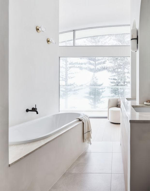 Bright, refined bathroom featuring a freestanding-style built-in bathtub beside expansive windows, a black wall-mounted bath tap, pale tiled flooring, white walls, and a light stone or concrete-look bath surround and vanity. A sculptural wall light and upholstered chair add understated luxury, while the large glazing provides an airy connection to the outdoors.