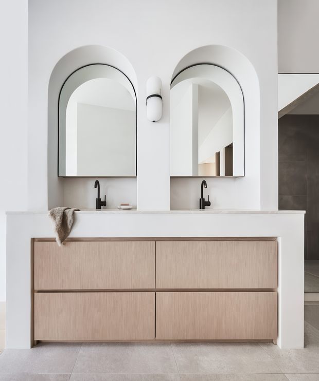 A bright, minimalist bathroom vanity with a wide double-sink basin, pale wood-grain cabinetry, matte black wall-mounted faucets, and two matching arched mirrors set into a white wall. A small wall sconce sits between the mirrors, while an open doorway and stair area are visible at the right edge.