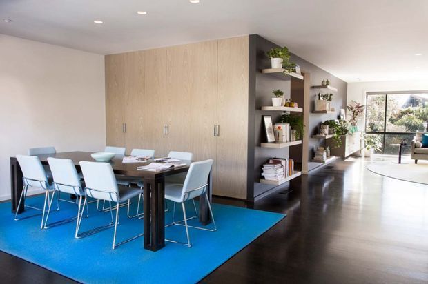 Open-plan dining and living area with a dark timber dining table and white upholstered dining chairs arranged on a vivid blue rug. A large light timber built-in storage wall and dark open shelving divide the dining zone from the bright living area, which opens toward full-height glazing and a balcony or exterior view. Recessed ceiling lights, indoor plants, books, and clean-lined joinery create a contemporary, polished interior.