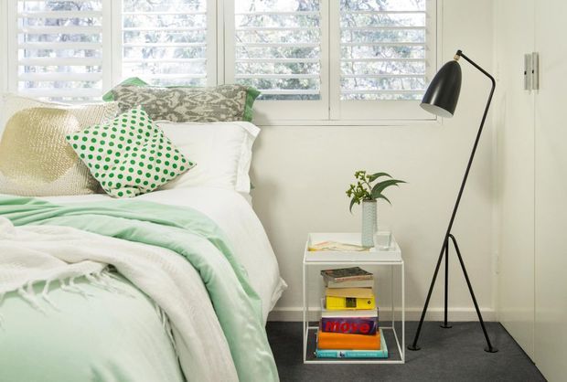 A bright, compact bedroom with a white bed dressed in layered white and pale mint bedding, patterned green-and-white cushions, and a small geometric side table stacked with books and topped with a plant. White plantation shutters cover the windows, while a tall black tripod floor lamp adds contrast. The room has a calm, fresh, contemporary feel with a dark low-pile carpet and light neutral walls.