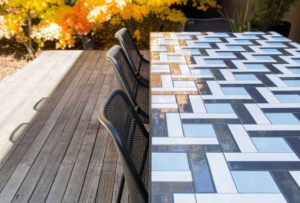 Outdoor dining area with a long rectangular tiled table on a timber deck. The tabletop features a bold geometric arrangement of rectangular tiles in white, pale blue-grey, and dark grey, paired with black mesh dining chairs. Autumn foliage and landscaped planting surround the deck, creating a contemporary garden setting.
