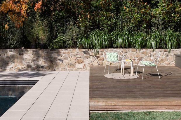 A landscaped outdoor poolside area with a dark timber deck beside a pale tiled pool surround. A low rough natural-stone retaining wall borders dense planting, while two light wire-frame chairs and a small table create a simple outdoor seating zone.