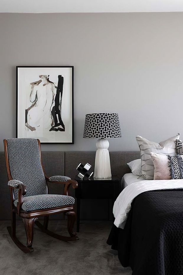 A sophisticated bedroom vignette featuring a grey upholstered wall or headboard, a dark upholstered bed with layered white and patterned bedding, a traditional timber rocking chair with black-and-white upholstery, and a black bedside table with a sculptural white lamp. Monochrome abstract artwork and patterned textiles add visual interest to the refined, high-contrast interior.