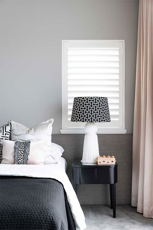 A stylish bedroom corner with a partially visible bed dressed in white linens and a dark charcoal quilt, patterned black-and-white cushions, a black bedside table, and a decorative table lamp. The room has soft grey walls, a white plantation-shutter window, a dark textured wall panel behind the bedside table, and full-length blush-beige curtains. The overall look is monochromatic, refined, and contemporary with graphic pattern accents.