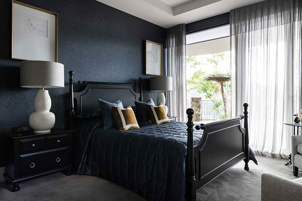 A dark, elegant bedroom with a black four-poster bed, deep navy patterned feature wallpaper, matching bedside tables and lamps, framed artwork, and layered charcoal bedding. Full-height sheer curtains soften the large window, creating a refined contrast between the moody interior and bright garden view outside.