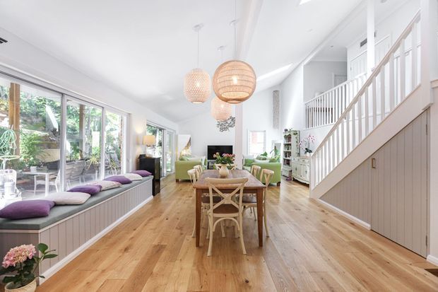 Bright open-plan living and dining area with white walls, extensive glazing, a built-in window seat, timber flooring, a timber dining table, pendant lights, and a staircase with white balustrades and vertical wall panelling. Lush greenery is visible outside through the glazing.