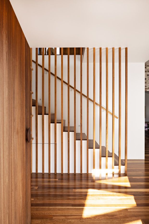 Interior view of a contemporary stairway with dark timber flooring and a timber-clad wall or door in the foreground. A straight staircase with dark wood treads and a simple wood handrail is screened by evenly spaced vertical timber balusters. White walls and ceiling create a bright, warm setting, with strong sunlight casting geometric patches across the floor.