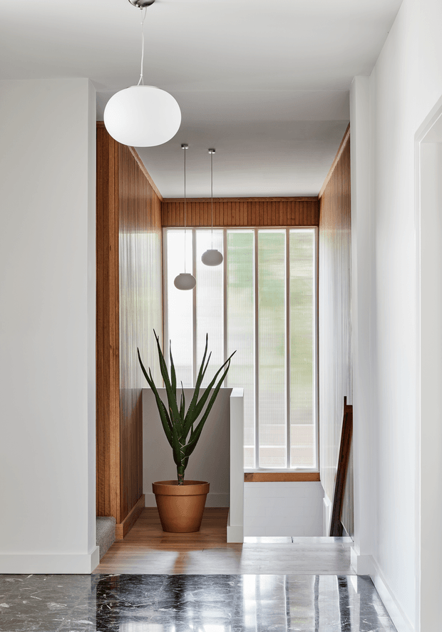A bright, narrow interior landing or hallway with a stair opening at the rear. The space features warm vertical timber wall panelling, tall translucent floor-to-ceiling glazing, polished dark stone flooring, white painted walls, and several suspended globe pendant lights. A large potted architectural plant provides a natural focal point.