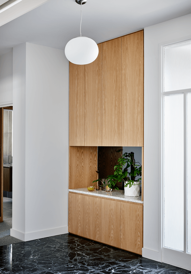 Bright contemporary interior featuring a full-height light timber built-in joinery wall with concealed upper and lower cabinetry and an open display niche. The niche contains indoor plants and a pale stone benchtop. A white globe pendant hangs from the smooth white ceiling, while dark veined stone flooring creates contrast. Tall windows with white vertical blinds bring in abundant natural light, and a partial adjacent kitchen or utility area is visible through the opening at left.