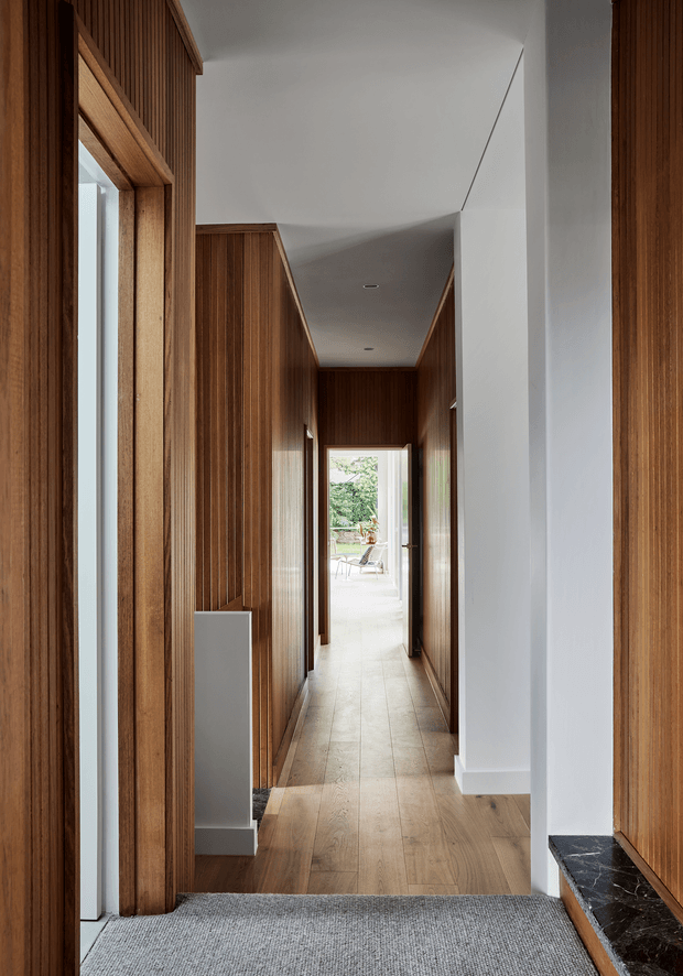 A long, narrow contemporary hallway lined with rich vertical timber wall panelling and timber-framed glazing. The corridor has warm timber flooring transitioning to a grey carpeted foreground, crisp white ceiling and walls, recessed downlights, and a dark stone window ledge at right. A bright outdoor seating area is visible at the far end, creating a strong axial perspective.