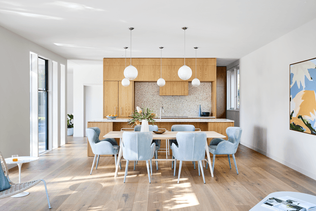 Bright contemporary open-plan dining and kitchen interior with a long timber dining table surrounded by upholstered light-blue chairs. The kitchen at the rear features warm timber cabinetry, a pale speckled stone or terrazzo-style splashback, and a wood-toned island bench. Large floor-to-ceiling windows bring in abundant natural light, while pendant globe lights and a colourful abstract artwork add visual accents. The room has wide-plank timber flooring and a calm, airy atmosphere.