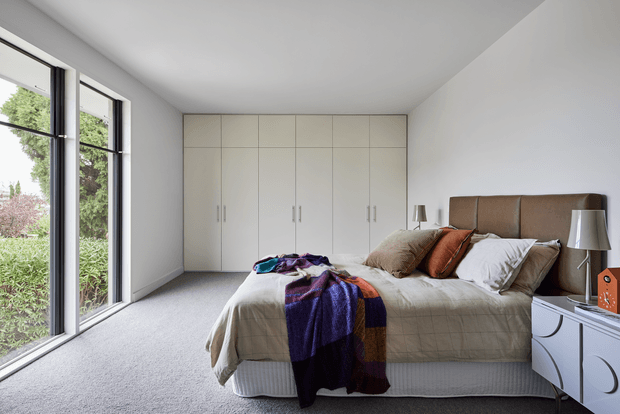 A bright, modern bedroom with a neatly made bed, upholstered brown headboard, white bedding, layered neutral and rust cushions, and a purple patterned throw. Full-height black-framed glazing opens to leafy garden views on the left, while a wall of pale built-in wardrobes provides storage at the rear. A curved-front white bedside cabinet and matching lamps complete the restrained contemporary interior.