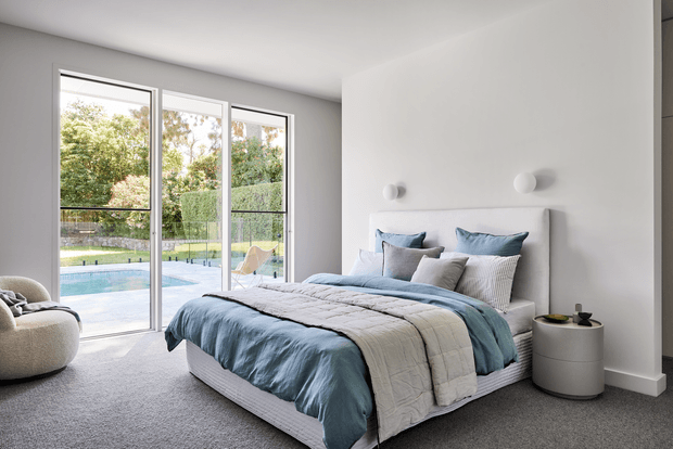 Bright contemporary bedroom with a large upholstered bed dressed in soft blue and grey bedding, grey carpet, white walls, minimalist bedside tables, and simple spherical wall lights. Full-height glazed doors open to a pool area and landscaped garden, bringing in abundant natural light.
