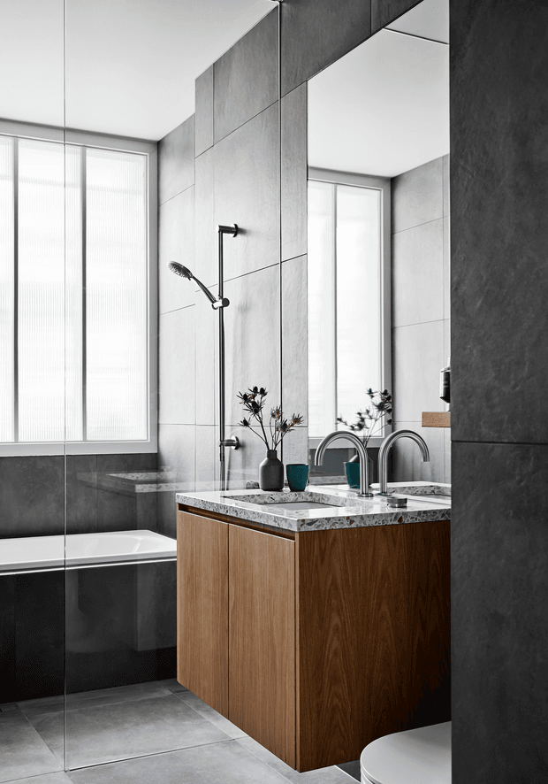 Contemporary bathroom with large-format grey wall and floor tiles, a freestanding-style white bathtub, frameless glass shower enclosure, mirrored wall, and a timber vanity with a speckled stone benchtop and twin curved basin taps. Soft daylight enters through tall textured windows, creating a refined monochromatic scheme with warm timber contrast.