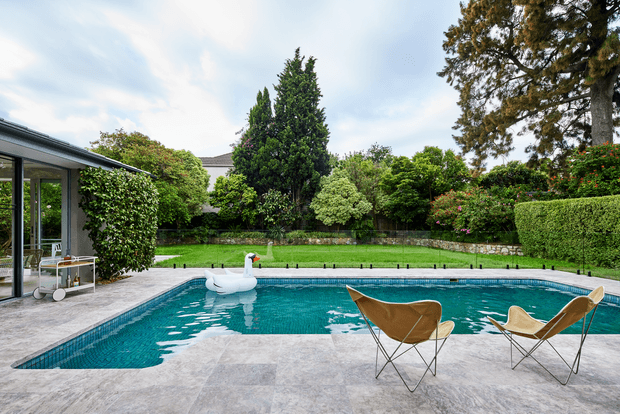 A landscaped residential backyard centered on a large rectangular swimming pool, with pale stone paving, two outdoor butterfly chairs, and a white inflatable swan. Dense mature garden planting, clipped hedges, a low stone retaining wall, and a glazed covered outdoor area frame the pool.