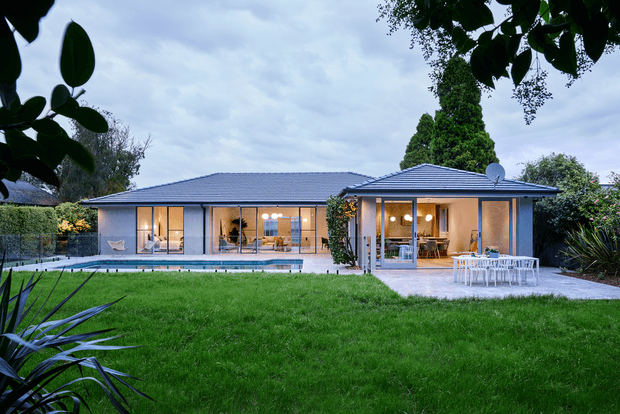 Single-storey contemporary suburban residence viewed from a landscaped backyard, with a broad low-pitched tiled roof, pale rendered walls, extensive floor-to-ceiling glazing, and warm interior lighting. A rectangular swimming pool runs along the house, with a paved outdoor dining terrace and manicured lawn in the foreground, surrounded by mature planting.