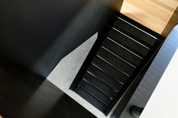 A top-down view of a contemporary stairway with dark textured stair treads and black side structure, bordered by pale grey flooring and dark walls. A light timber-clad landing or ceiling element is visible at the upper end, creating a strong monochrome contrast with the timber accent.