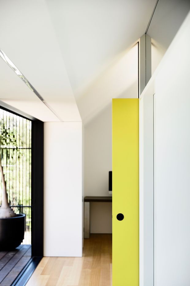 A bright, minimalist interior passage viewed toward a compact work area, with white walls and ceiling, pale timber flooring, a vivid lime-yellow pivot door or screen with a black circular handle, and a dark-framed glazed opening with vertical screening on the left. The space has clean geometric lines, concealed detailing, and strong contrast between white surfaces, black framing, natural timber, and the yellow feature element.
