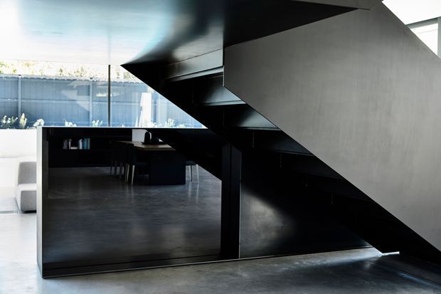 A contemporary interior focused on a dramatic black steel stair with a solid sculptural side wall and open-tread construction. The stair rises over a dark built-in workspace or dining area, with a large reflective panel beneath it. The surrounding space has polished grey concrete flooring, dark joinery, expansive glazing, and strong geometric lines.