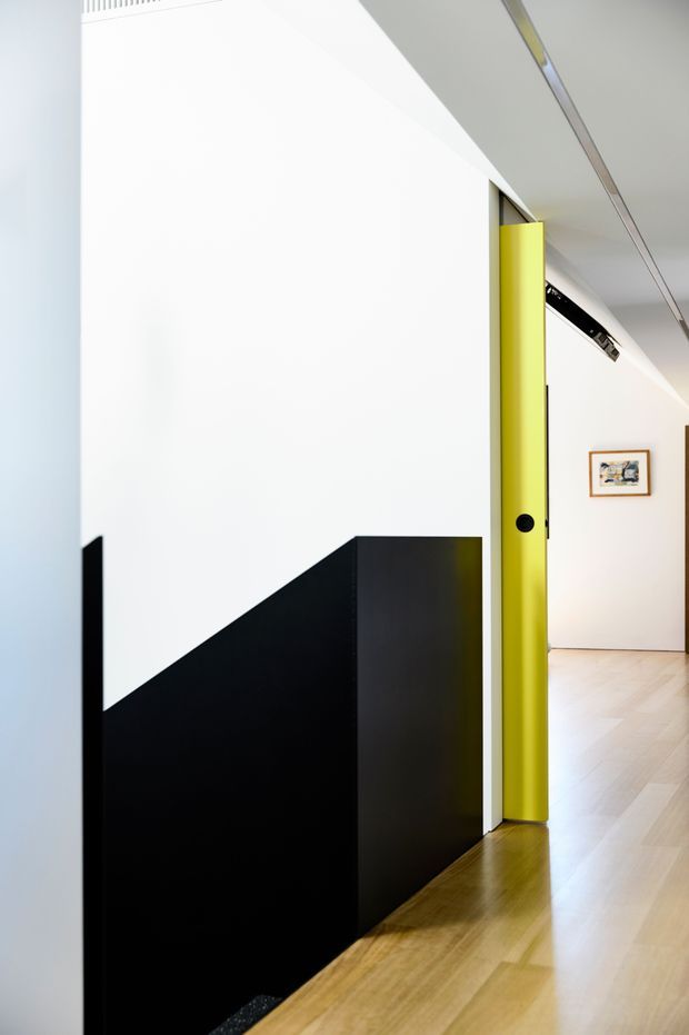 A minimalist interior hallway or entry passage with a striking geometric black-and-white wall and built-in black joinery or stair enclosure. A vivid lime-yellow pivot or sliding door creates a strong accent beside the opening into a bright gallery-like corridor. The space has pale timber parquet flooring, white walls and ceiling, slim linear ceiling detailing, and framed artwork in the adjoining area.