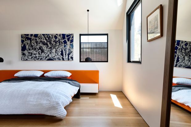 A bright contemporary bedroom with a low bed featuring a bold orange headboard and white bedding. The room has pale timber flooring, white walls, black-framed windows, a simple black pendant light, abstract blue-and-white wall art, and a full-height mirrored wardrobe or door reflecting the bed.