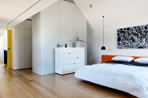 Contemporary bedroom with a low-profile bed, vivid orange upholstered headboard, white textured bedding, and a monochrome abstract artwork. The room features pale timber flooring, white built-in cabinetry and wardrobes, a white chest of drawers, a slim black pendant light, and a bright yellow door accent. Clean-lined, uncluttered interior with strong geometric forms.