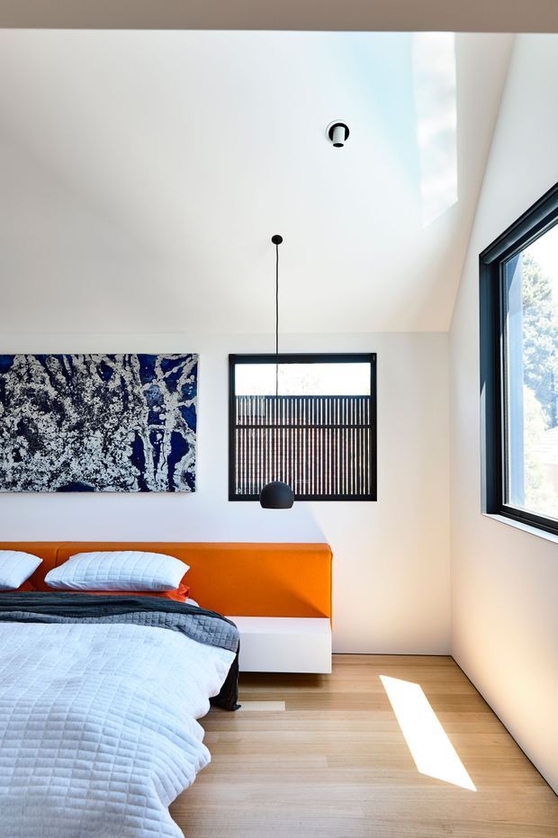 Bright contemporary bedroom with a low platform bed, vivid orange upholstered headboard, pale quilted bedding, and a dark blue abstract artwork. The room has white walls and ceiling, warm timber flooring, black-framed windows, a small black pendant light, and strong natural light entering through the glazing. A slatted screen is visible outside the central window.