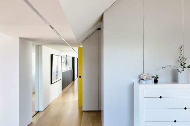 A bright, minimalist interior hallway with pale timber flooring, white walls and ceilings, and extensive flush built-in cabinetry. A vivid yellow door or wall panel provides a strong accent at the end of the corridor. The foreground includes a simple white chest of drawers with understated black pulls and sparse decorative branches. Recessed linear lighting and clean, handleless joinery create a streamlined contemporary appearance.