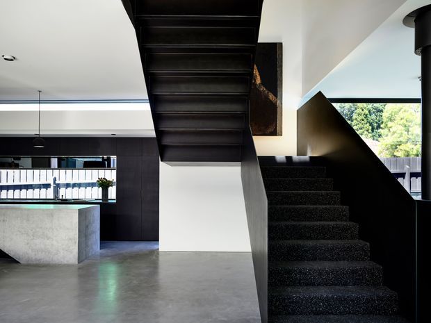 A contemporary open-plan interior centred on a dark sculptural staircase with black carpeted steps and a dark timber or metal soffit. The staircase divides a minimalist kitchen and living area, with a concrete island, dark cabinetry, polished grey concrete flooring, white walls, linear lighting, and large windows overlooking greenery. The space has strong geometric forms and a high-contrast black, white, and grey palette.
