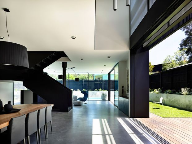 A contemporary open-plan residential interior with a dining area in the foreground, a bold black sculptural staircase, polished concrete flooring, and a double-height glazed living area opening to a timber deck and landscaped courtyard. Strong daylight enters through expansive floor-to-ceiling glazing, creating a bright connection between the interior and outdoor space.