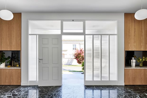 Bright, refined residential entryway viewed from inside, featuring a grey paneled front door with sidelights and transom glazing, full-height fluted or textured glass panels, warm timber built-in cabinetry with dark recessed niches on both sides, and a highly polished dark marble-look floor. Minimal white pendant lights and strong symmetry create a contemporary, elegant foyer.