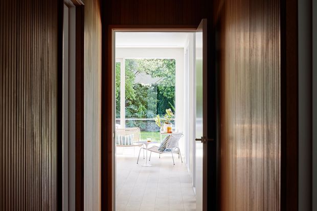 A dark, narrow interior passage framed by rich timber and vertically fluted wall panelling leads through an open doorway into a bright glazed sitting area. The adjoining room has pale flooring, light furniture, and extensive views toward a leafy garden, creating a strong contrast between the moody corridor and airy room beyond.