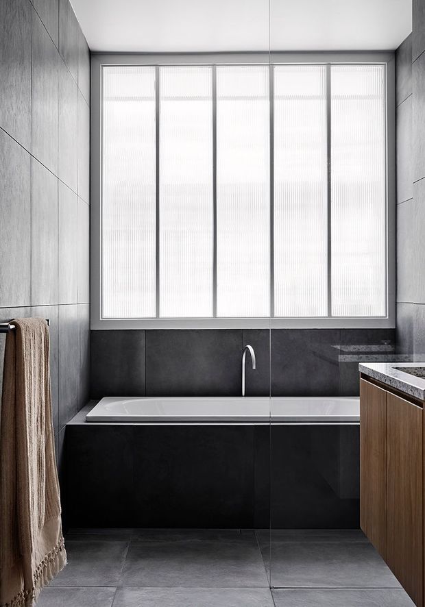 A compact, contemporary bathroom featuring a built-in white bathtub beneath a large multi-panel translucent window. Dark grey full-height wall and bath surrounds create a monochromatic, minimalist setting, with large-format grey floor tiles, a slim curved chrome tap, and a timber vanity with a pale stone benchtop at right. A neutral towel hangs at the left edge.