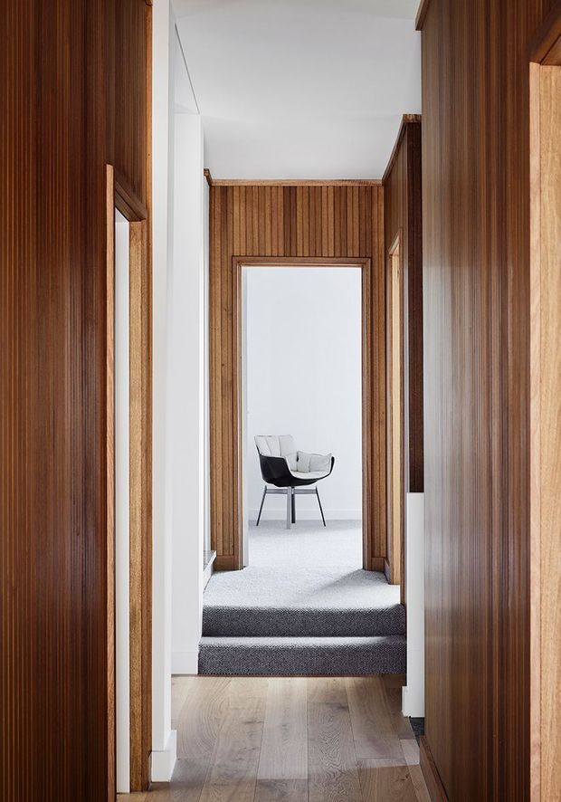 A narrow, refined interior hallway lined with vertical dark timber wall panelling and matching timber door frames. The corridor transitions from a pale timber floor to charcoal carpeted steps and landing, leading toward a bright room containing a sculptural black-and-white chair. White ceilings and wall sections provide contrast, while recessed doorways and crisp detailing create a carefully framed view through the home.