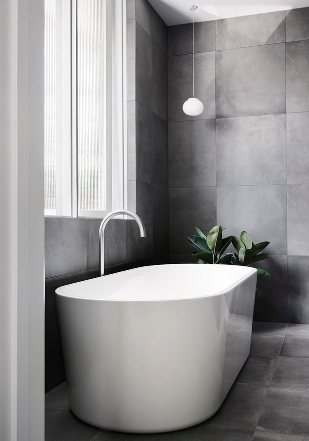 A refined contemporary bathroom featuring a freestanding white oval bathtub, dark grey large-format tiled walls and floor, a curved chrome bath tap, a minimalist globe pendant light, and a leafy indoor plant. Tall white-framed vertical windows bring in natural light, creating a cool, calm and luxurious atmosphere.