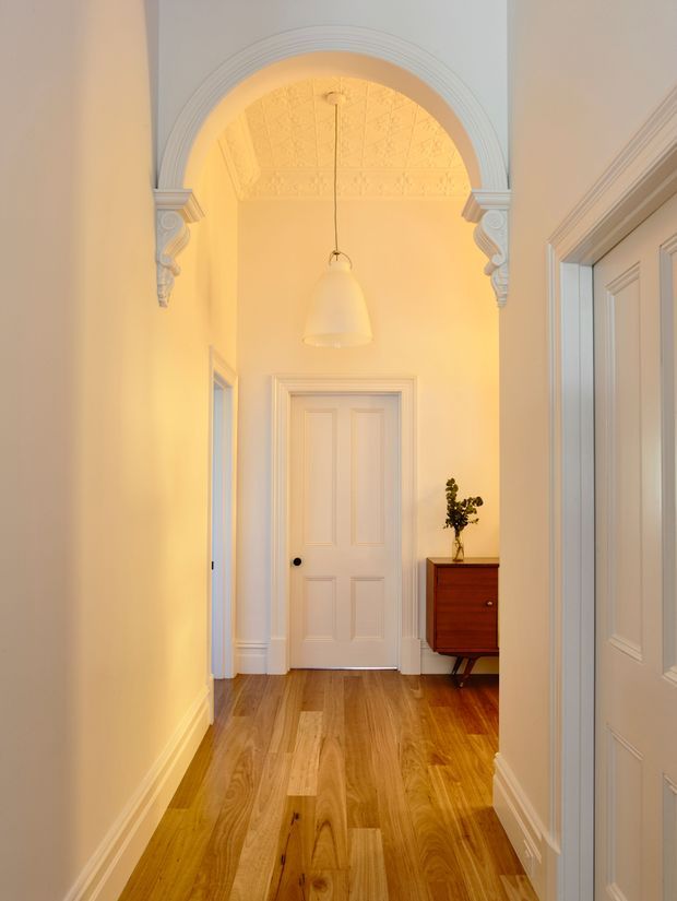 A warm, refined residential hallway viewed through an ornate arched opening. The corridor features honey-toned timber flooring, pale painted walls, detailed white moulded architraves and cornices, decorative classical brackets, a patterned pressed ceiling, paneled interior doors, and a small dark timber chest with a vase. A simple pendant light hangs centrally, creating a softly illuminated atmosphere.