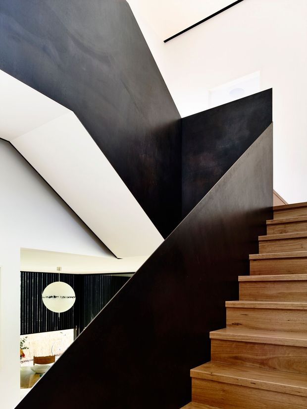 Interior architectural photograph focused on a contemporary stairway. Broad angular black and dark metal-toned balustrade and wall planes create a dramatic geometric composition, contrasted with warm timber stair treads. White walls and ceiling, slim black linear detailing, and a glimpse of a lower-level room with dark vertical wall panelling are visible.