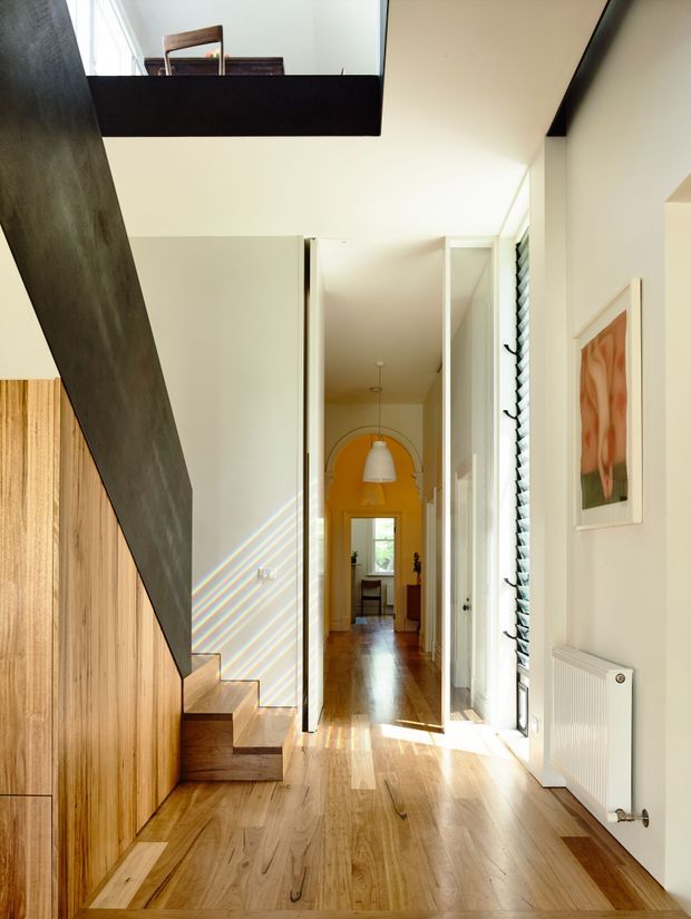 Bright contemporary hallway with warm timber flooring and matching timber stair and cabinetry elements. A dark sculptural stair stringer frames the left side, while white walls, a tall glazed opening with louvred shading, and a white radiator line the corridor. The hallway extends toward a colourful yellow-orange arched passage with pendant lighting and visible dining or living space beyond. An upper-level opening and refined minimalist detailing create a strong vertical connection and airy feel.
