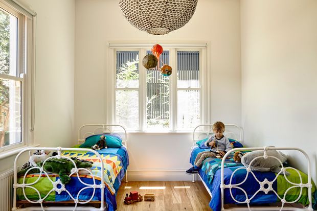 Bright shared children's bedroom with two white metal-framed beds, colourful patterned quilts, toys, and a woven pendant light. Tall windows provide abundant natural light, while a warm timber floor and pale walls create an airy, playful room.
